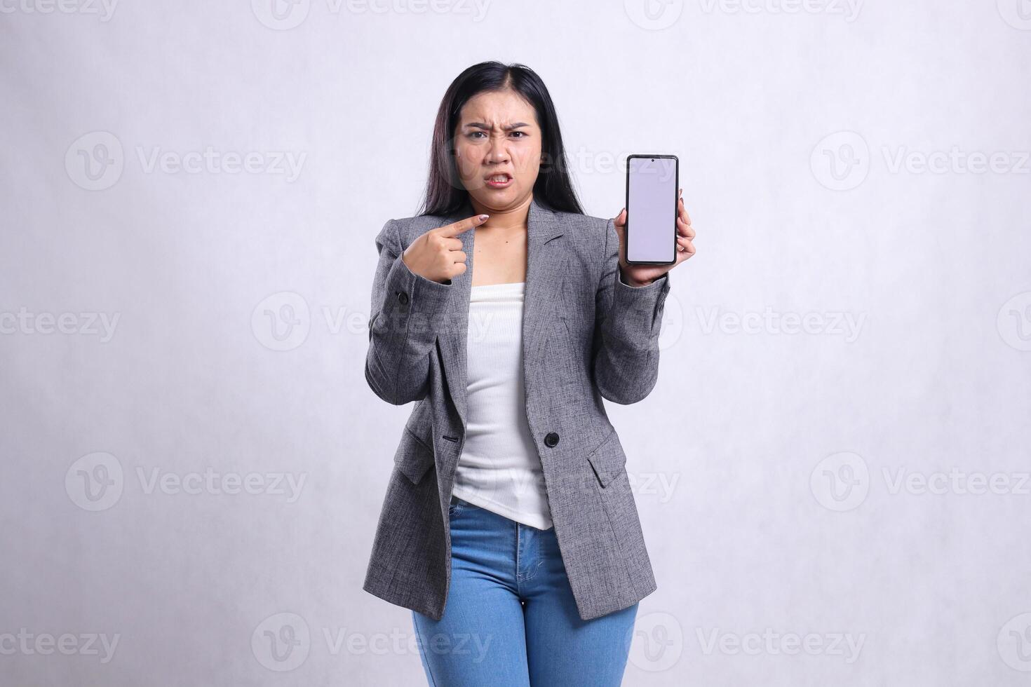 beautiful mature office woman asia gesture angry to camera pointing holding showing cellphone screen wearing grey suit isolated on white background. for technology, lifestyle, promotion content photo
