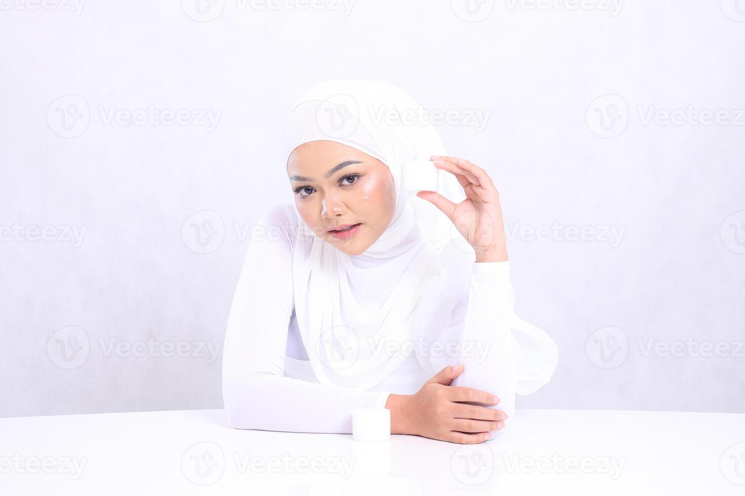 Beautiful young hijab woman expression looking down at camera hand holding cosmetic cream on left side of face for daily skin care routine needs on white background. Facial treatment, Perfect skin photo