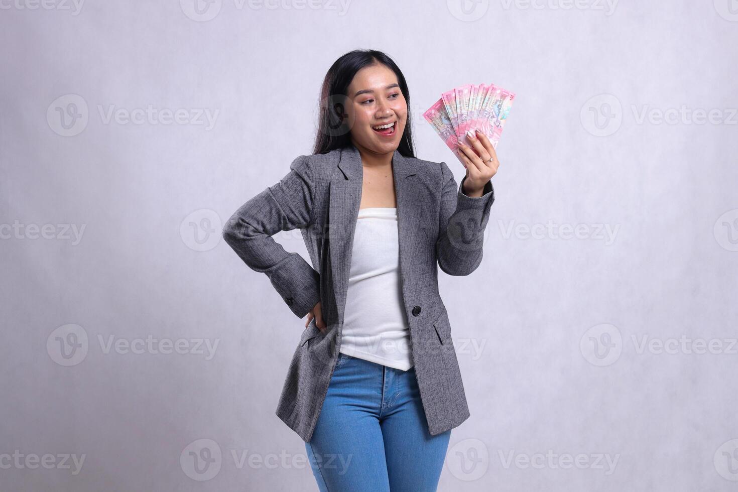expression of beautiful office female asia happy candid hand holding rupiah money and waist wearing gray suit isolated on white background. for giveaway content, lifestyle, prize, event, competition photo