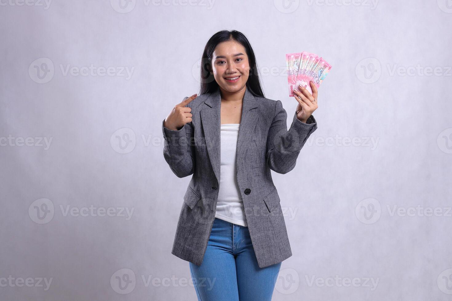 gesture beautiful young office female smile wide hand pointing and hold rupiah money wearing gray suit isolated on white background. for giveaway content, lifestyle, prize, event, competition photo