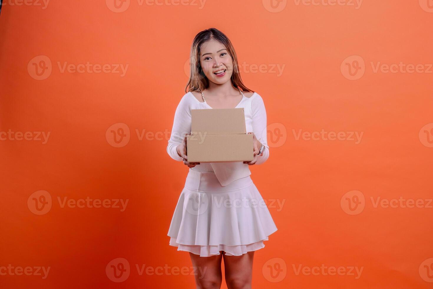 Charming young beautiful woman in white top cheerful to camera both hands holding 2 plain cardboard boxes, standing on orange background. for shipping concept, gift, expedition, service, package photo