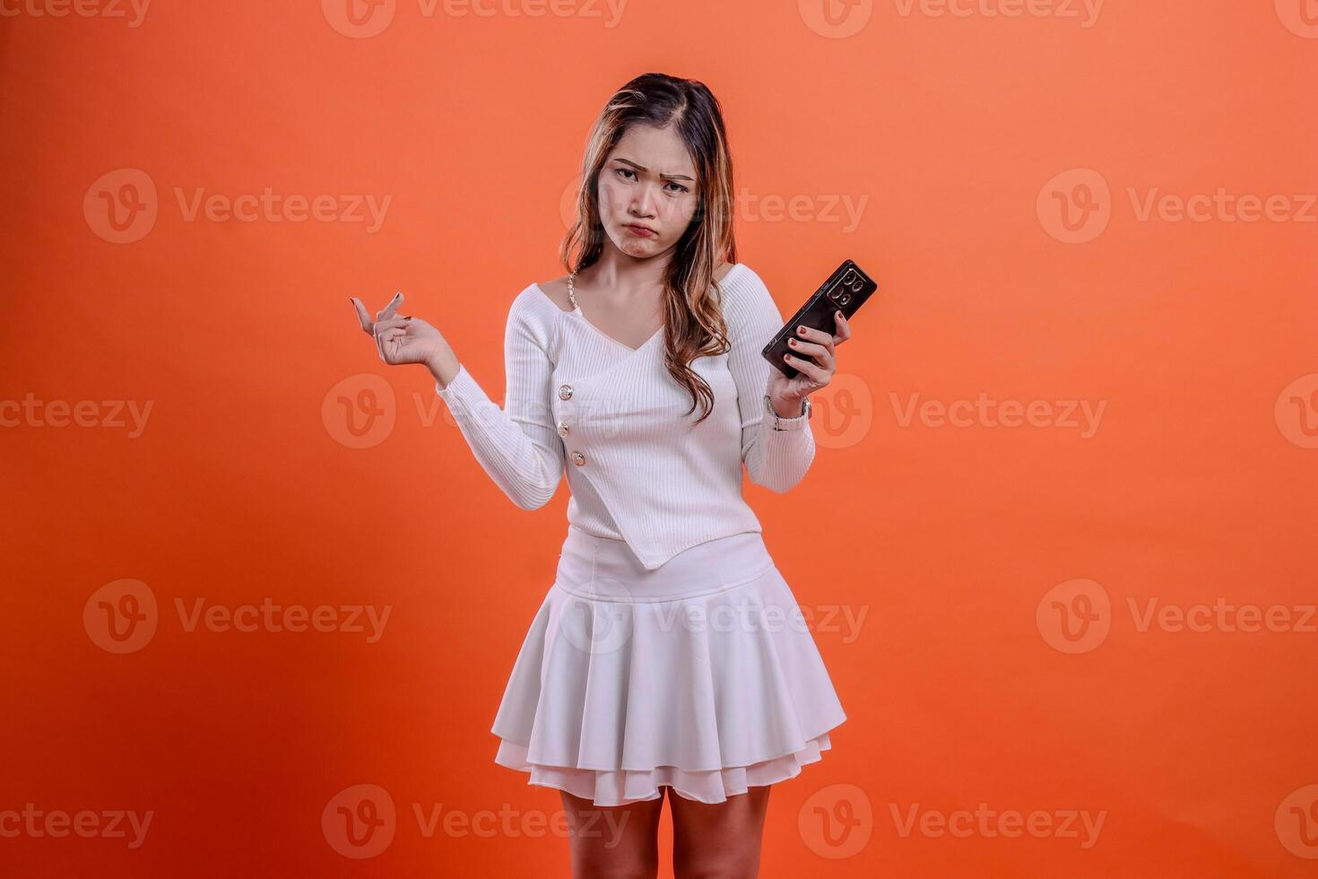 beautiful young ladies gesture in white top standing pouting to camera holding mobile phone, cellphone and hand pointing to right isolated orange background. for technology, promotion, lifestyle photo