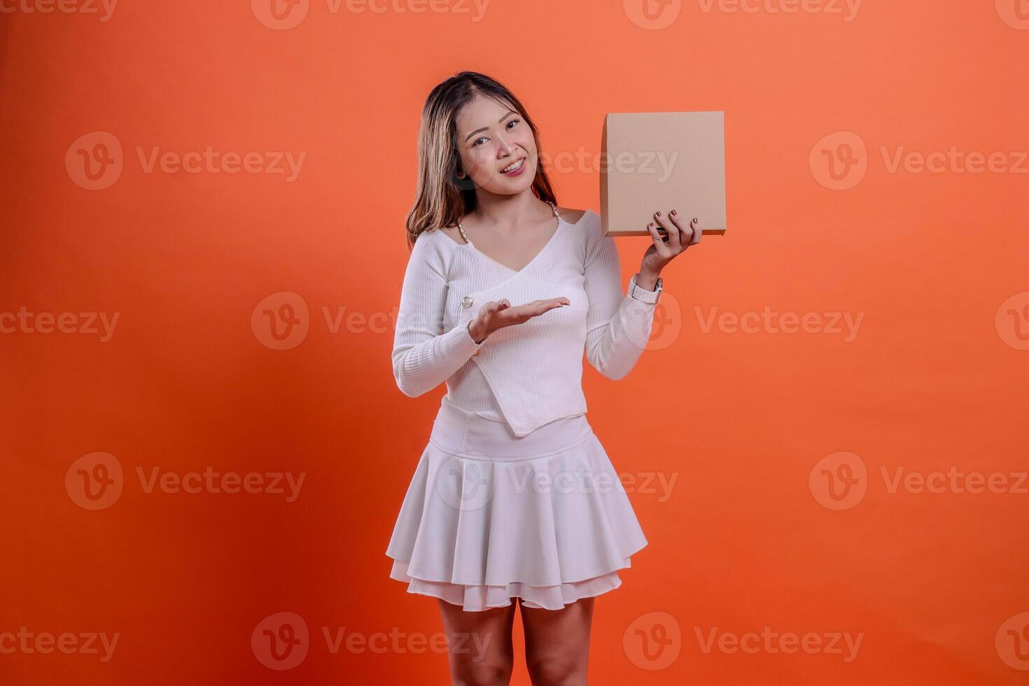 gesture of young asian girl in white top cheerful to camera hand carrying plain cardboard box while presenting, standing on orange background. for delivery concept, gift, expedition, service, package photo