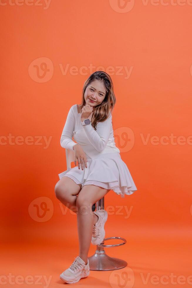 full body teenage girl white sweater cute smile slanted right hand hold chin chair sitting on hydraulic chair, standing on orange background. for fashion, lifestyle, beauty concept photo