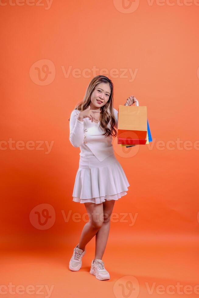 full body charm asian girl white top standing cheerful looking at camera hand lifting shopping bag forward while pointing at it orange background. for fashion concept, advertisement, lifestyle photo