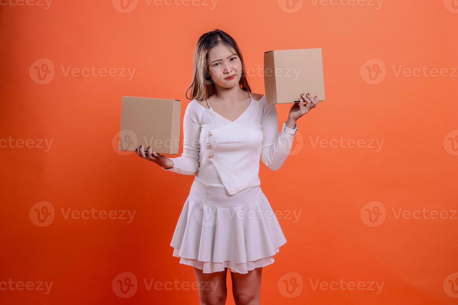 beautiful asian woman in white top pouting at camera both hands lifting plain cardboard box up and down, standing on orange background. for delivery concept, gift, expedition, service, package photo