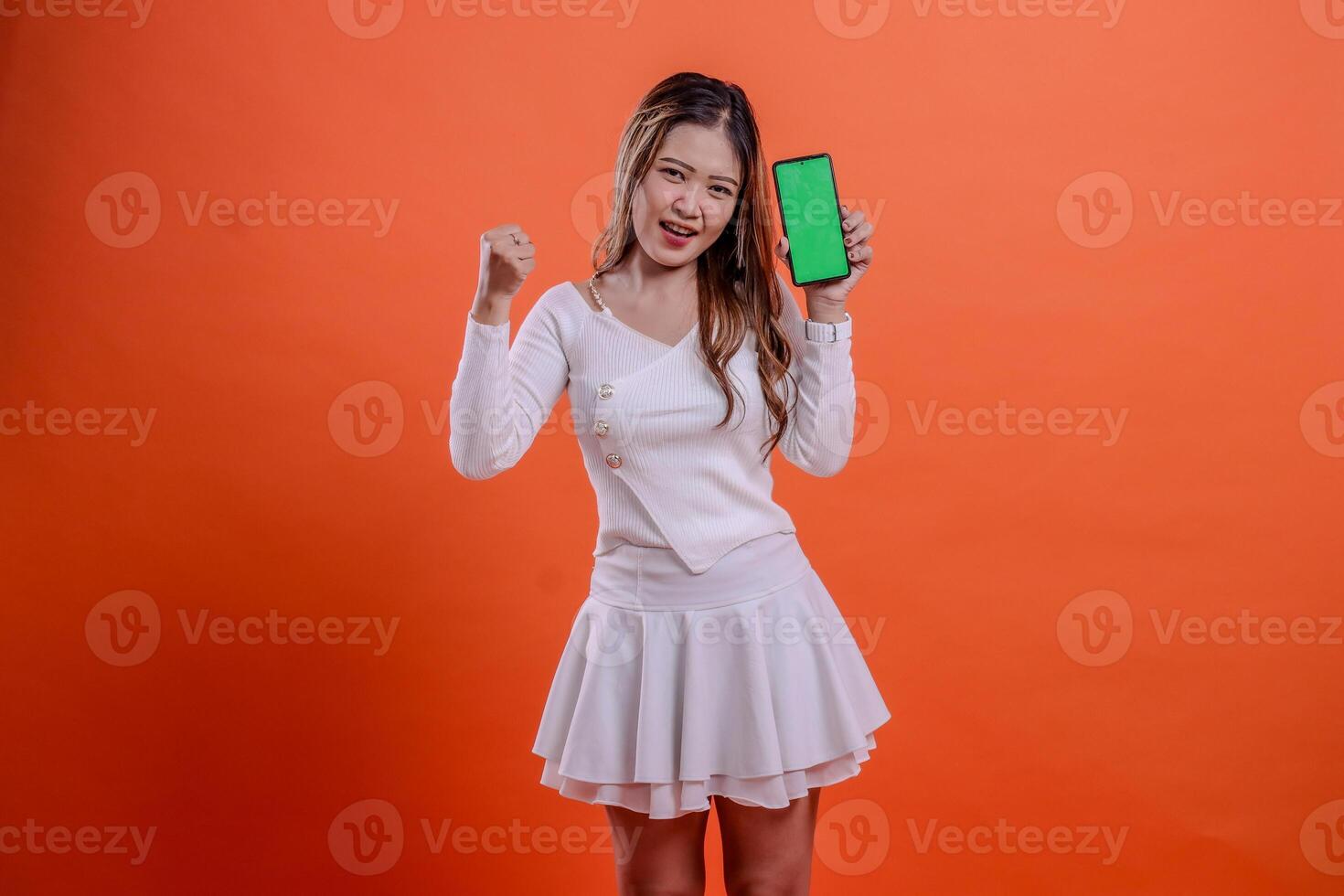 woman white top standing cheerful to camera both hands hold mobile phone screen, cellphone while clenched to the side isolated orange background. for technology, business, promotion, lifestyle concept photo
