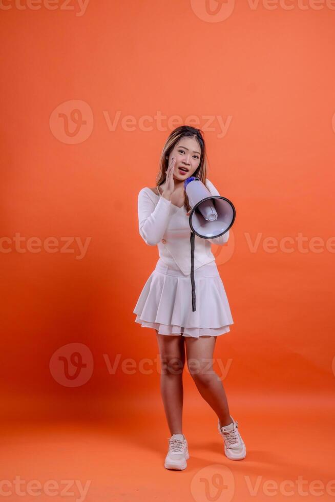 full length asian female white top standing to camera screaming angry hand holding megaphone loudspeaker while focusing sound isolation orange background. for technology concept, advertising photo
