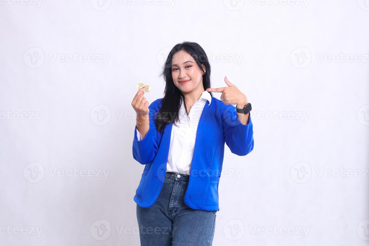 gesture of beautiful adult office woman wearing blue formal suit standing smiling to camera holding bitcoin while pointing at it isolated white background. for transaction content, investment photo