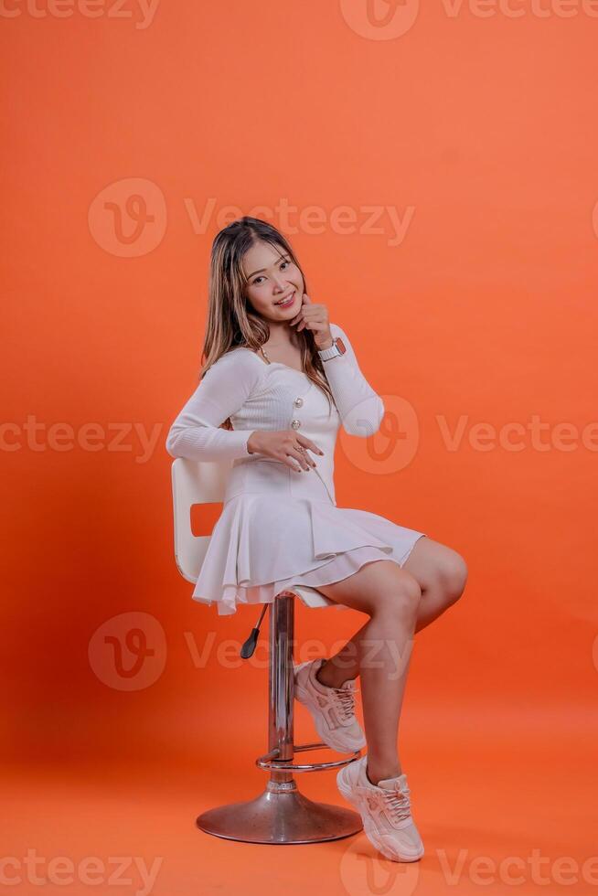 full length young asian girl cheerful white sweater sender on chair hold chin slanted left sitting on hydraulic chair, standing on orange background. for fashion, lifestyle, beauty concept photo