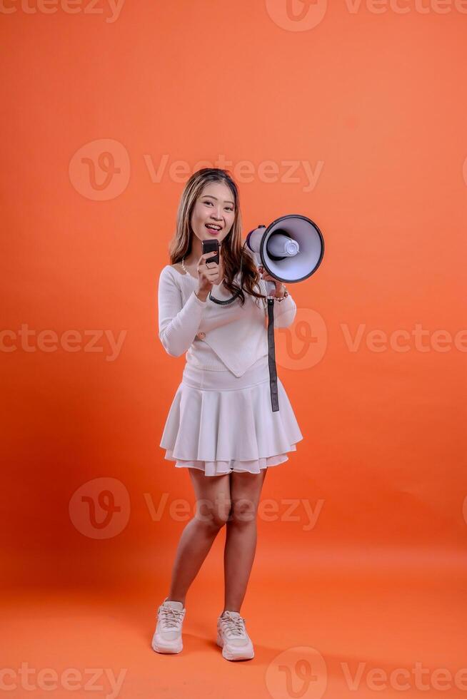 full body beautiful woman white top standing to camera screaming happily holding megaphone loudspeaker forward and mic audio isolation orange background. for technology concept, advertising, broadcast photo