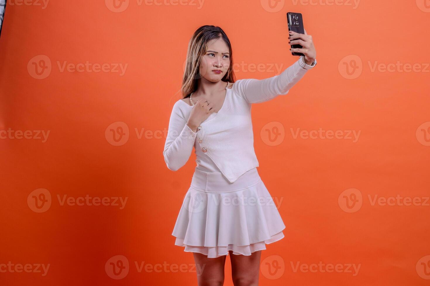indonesia woman in white sweater pouting hands on chest call using smartphone camera, mobile phone, cellphone, standing on orange background. for technology, lifestyle, business, service concept photo