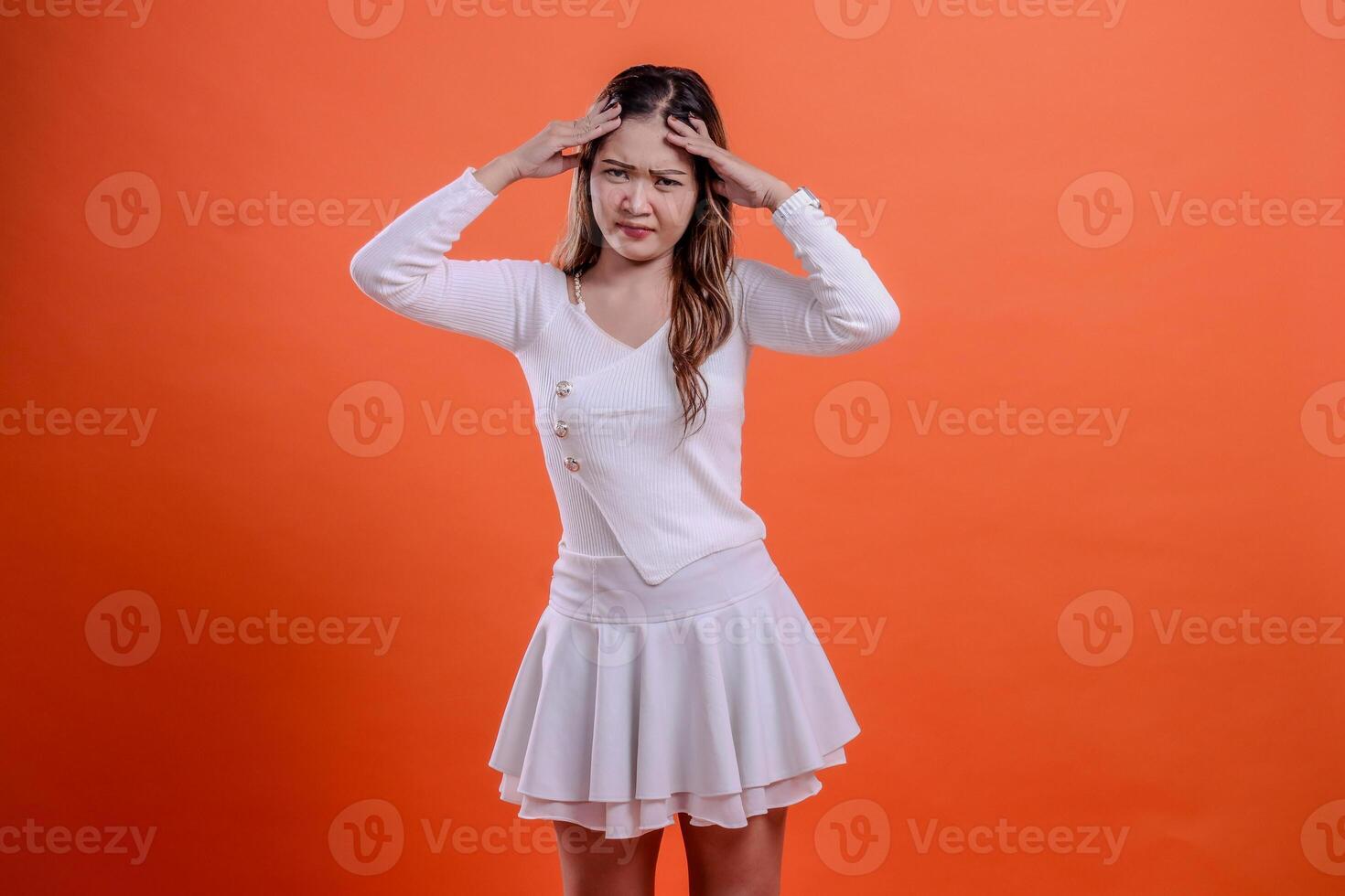 portrait of beautiful young asian woman in white top standing sad dizzy to camera both hands hold head isolated orange background. for health concept, advertising, science, commercial photo