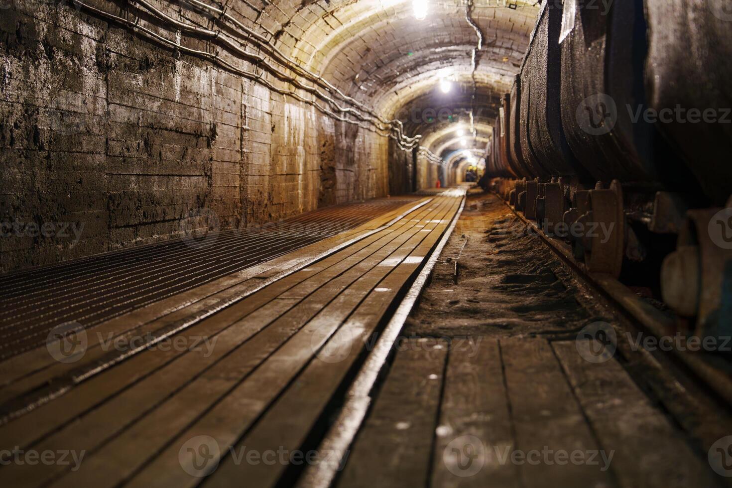 The image showcases an old, rusty mine tunnel, featuring weathered steel tracks that lead into darkness. Vintage mining carts are scattered throughout, evoking a sense of history and adventure. photo
