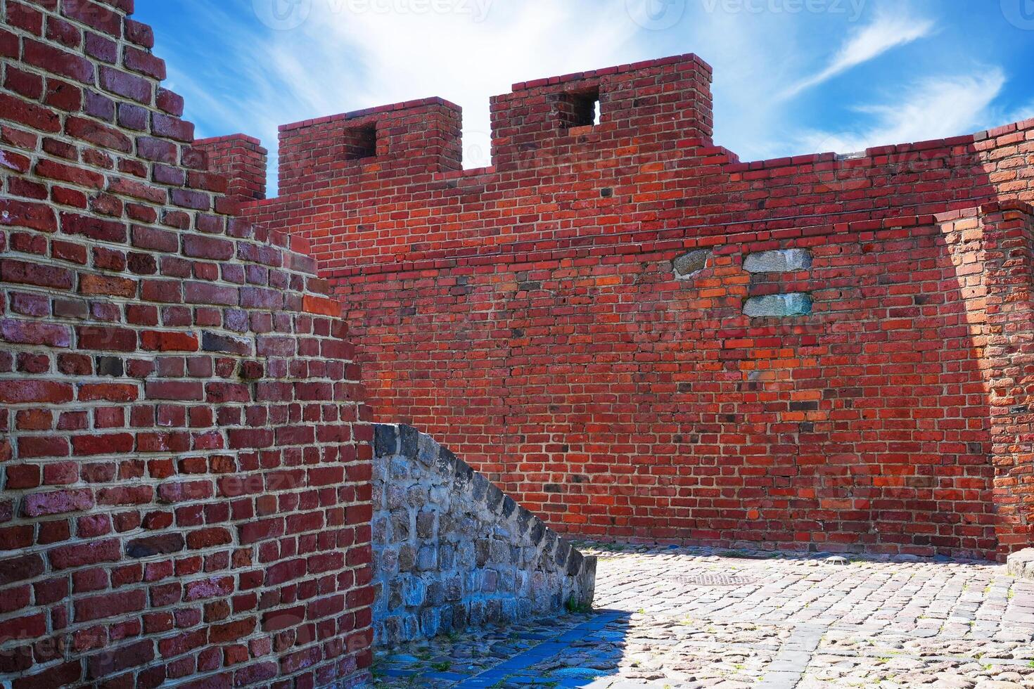 A historic red brick fortress wall with two turrets stands under a clear blue sky. A charming cobblestone street leads into the distance, featuring a hole in the larger turret. photo