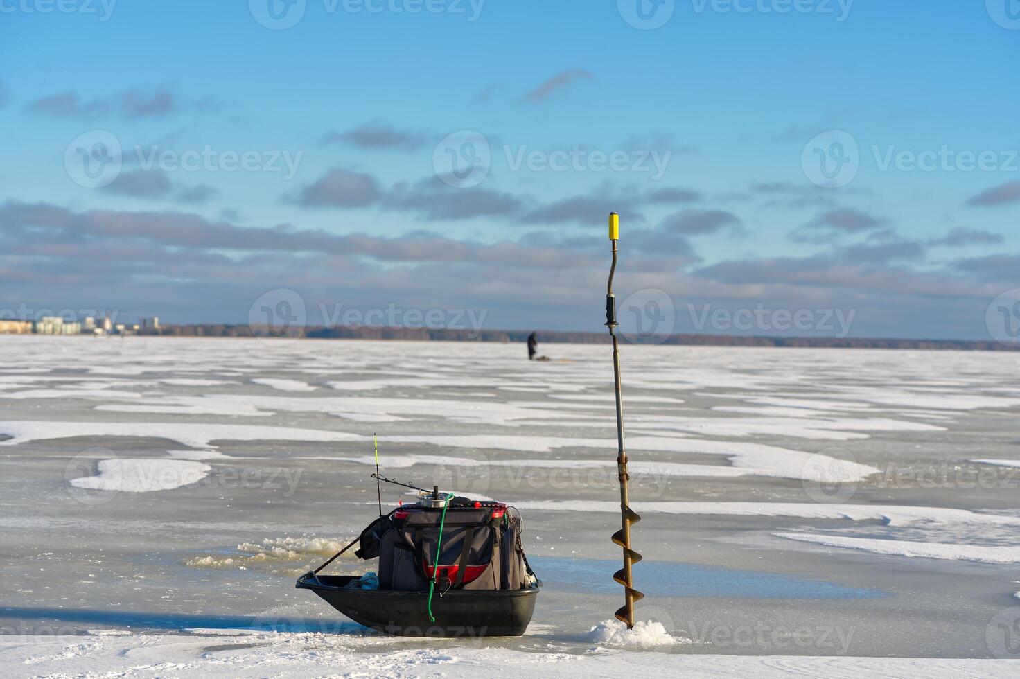 An ice fishing hole is drilled in the ice with a fishing rod in it. A sled with a tackle box and gear is nearby, while a person is seen ice fishing in the distance. photo