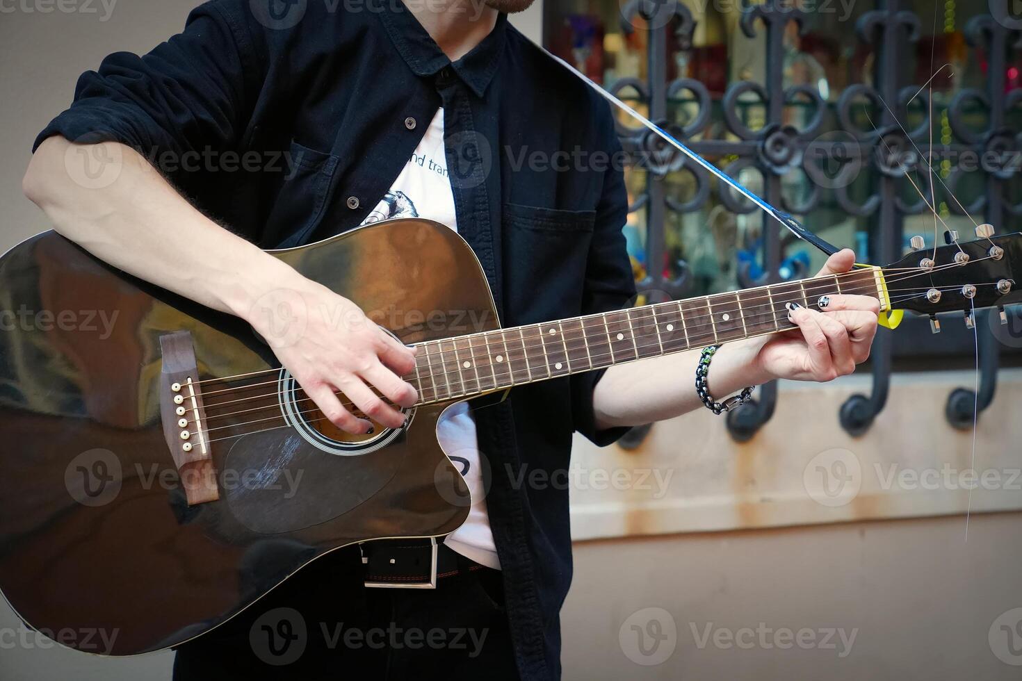 A young male with a beard and black hair plays an acoustic guitar outdoors. The scene features a reflective surface with artistic patterns, contributing to a serene atmosphere. photo