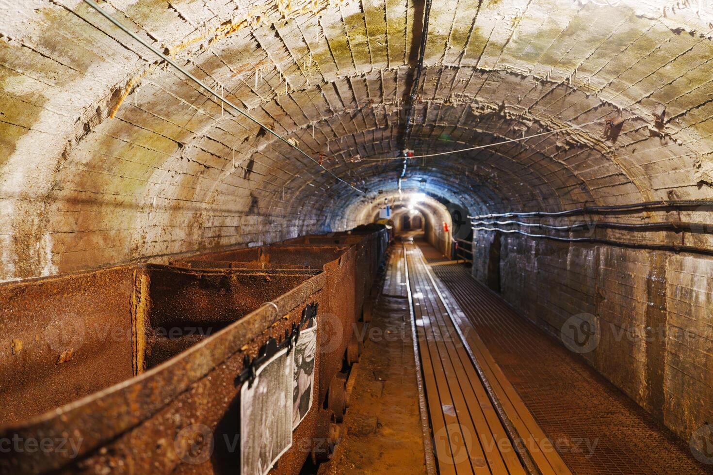 The image captures a vintage mining cart resting on rusty tracks in a dark mine tunnel. The atmosphere is mysterious, with a brick ceiling and shadows creating an eerie ambiance, gold mine photo