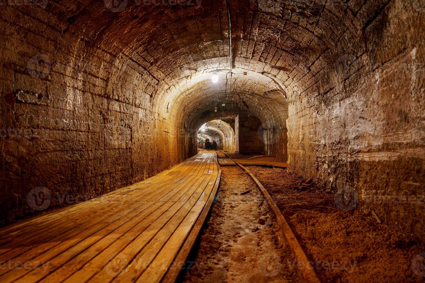 A dimly lit tunnel features a worn wooden walkway and rusty railroad tracks. The rough stone walls add to the dark, mysterious atmosphere of this abandoned passage, cooper mine photo