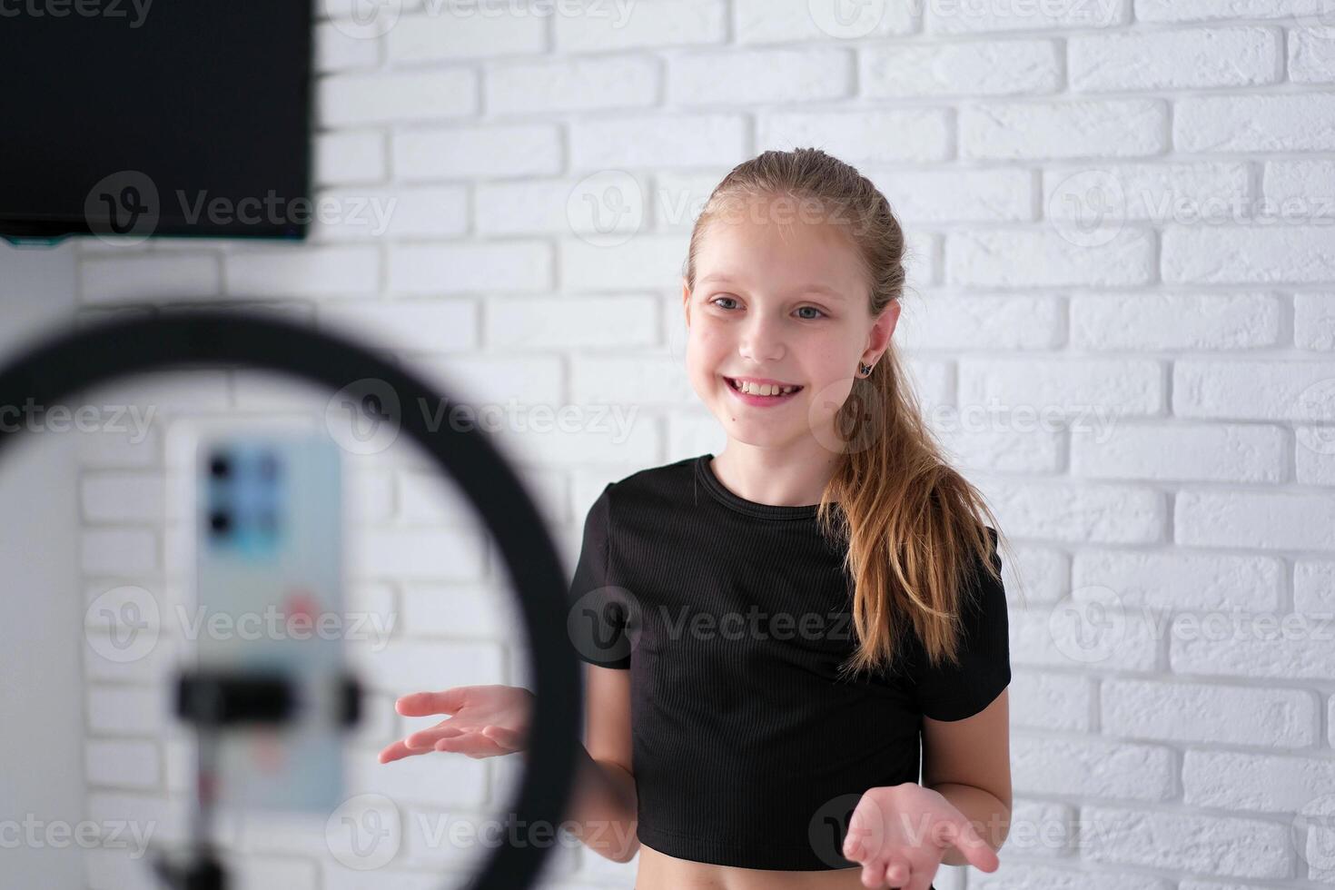 Beautiful teenage girl posing in front of a phone on a tripod. Child blogger shoots in front of camera and ring lamp photo