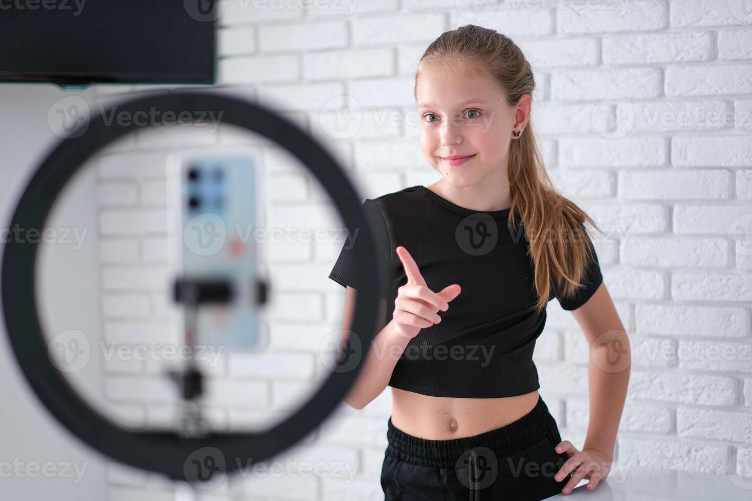 Beautiful teenage girl posing in front of a phone on a tripod. Child blogger shoots in front of camera and ring lamp photo