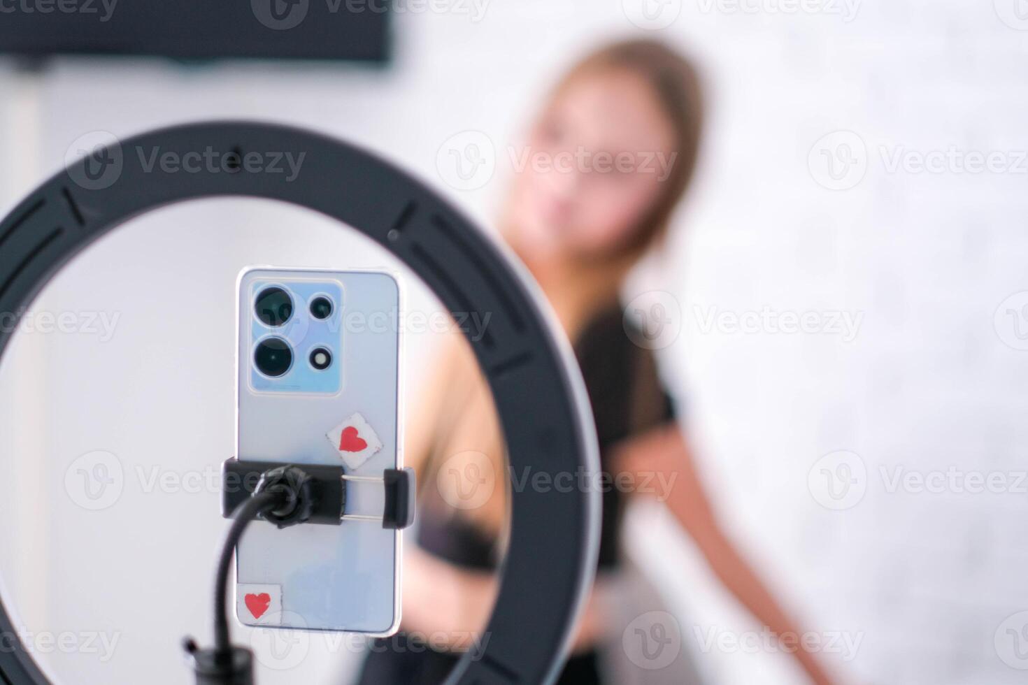 A teenage girl poses in front of a phone on a tripod. A child blogger shoots a blog. Focus on the ring lamp and telephone. photo
