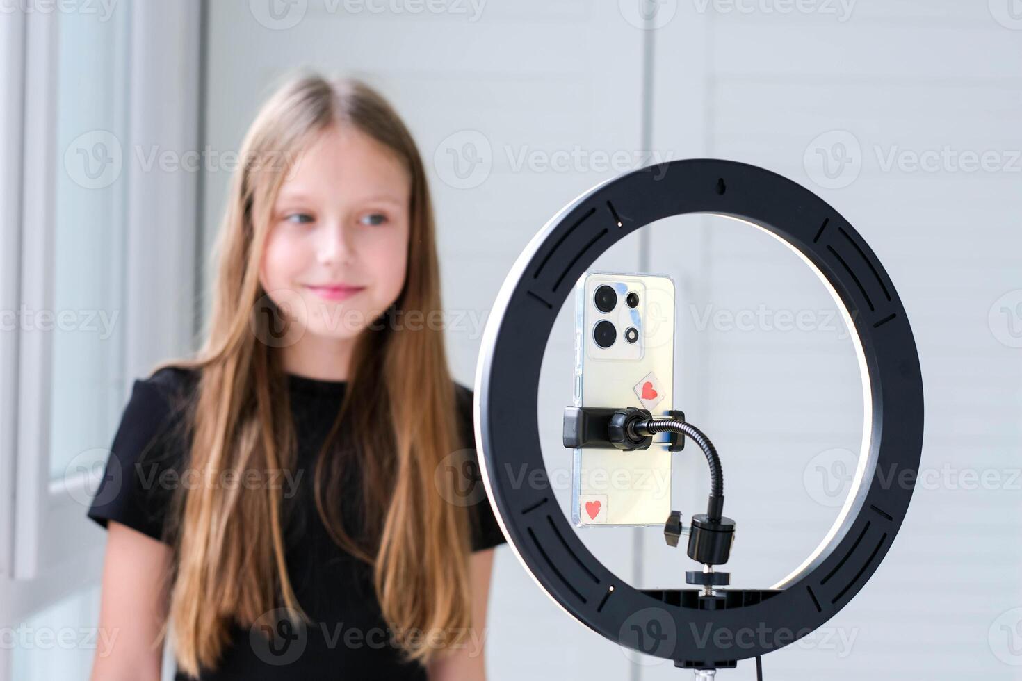 Beautiful teenage girl posing in front of a phone on a tripod. Child blogger shoots in front of camera and ring lamp photo