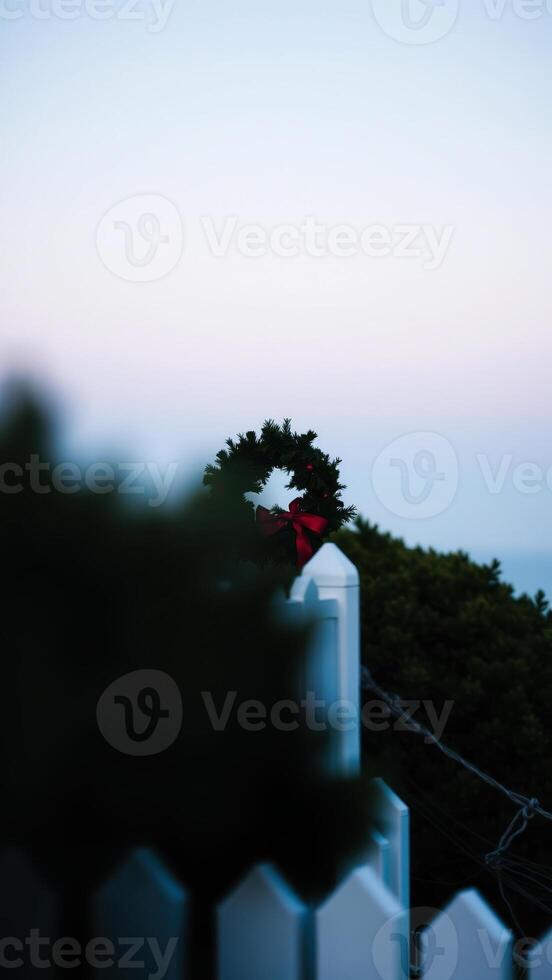 festive evergreen wreath with a red bow atop a white picket fence post photo