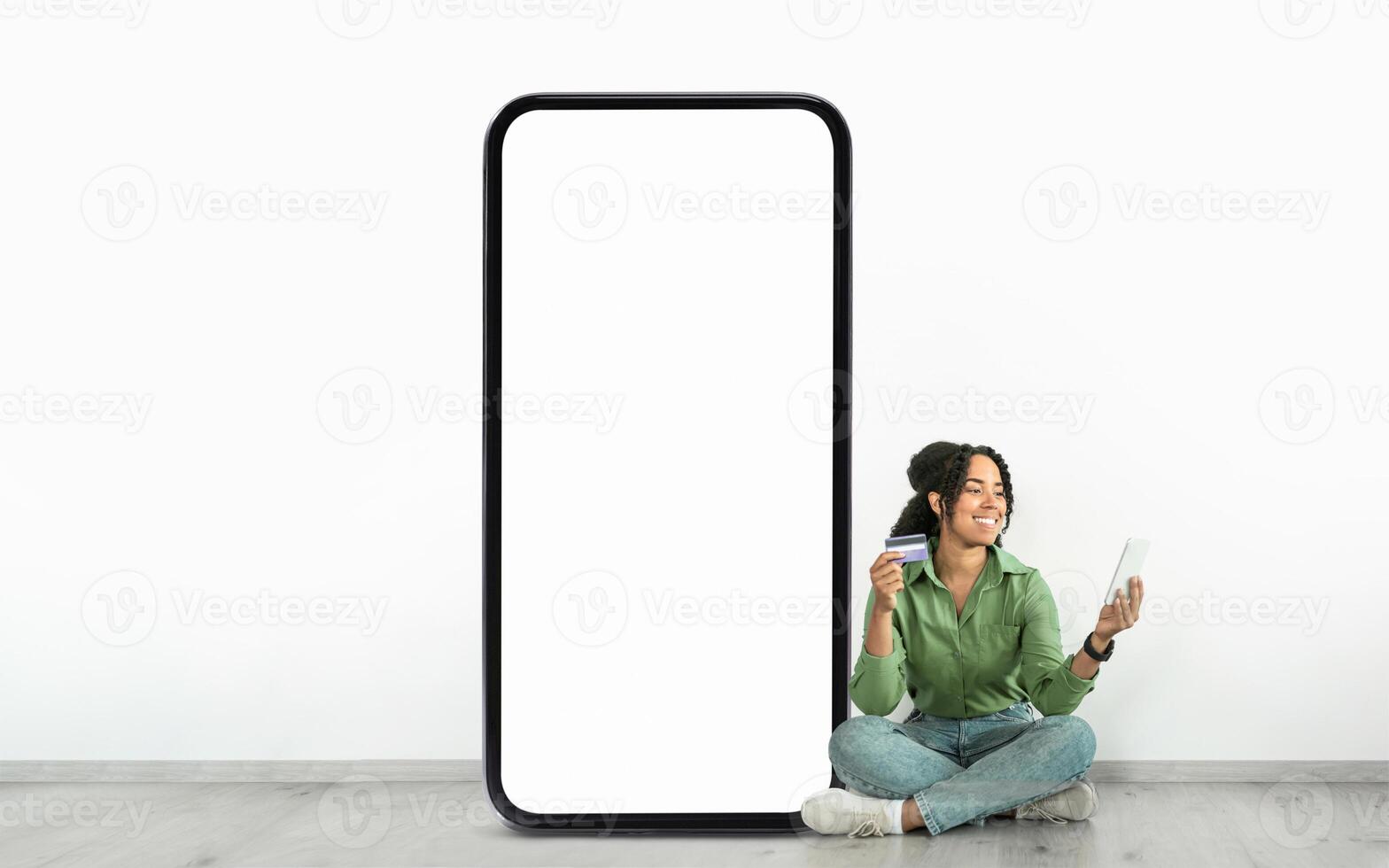 A woman is sitting on the floor, smiling as she holds a credit card and a tablet, next to a large blank smartphone display. The setting suggests a relaxed digital shopping experience. photo