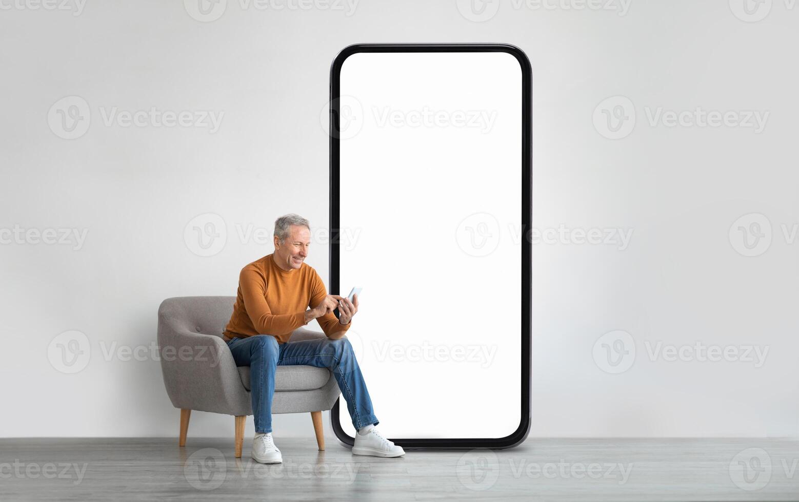 An older gentleman in a brown sweater sits comfortably in a gray chair, engaged with his smartphone, beside a large blank smartphone display in a minimalistic room. photo