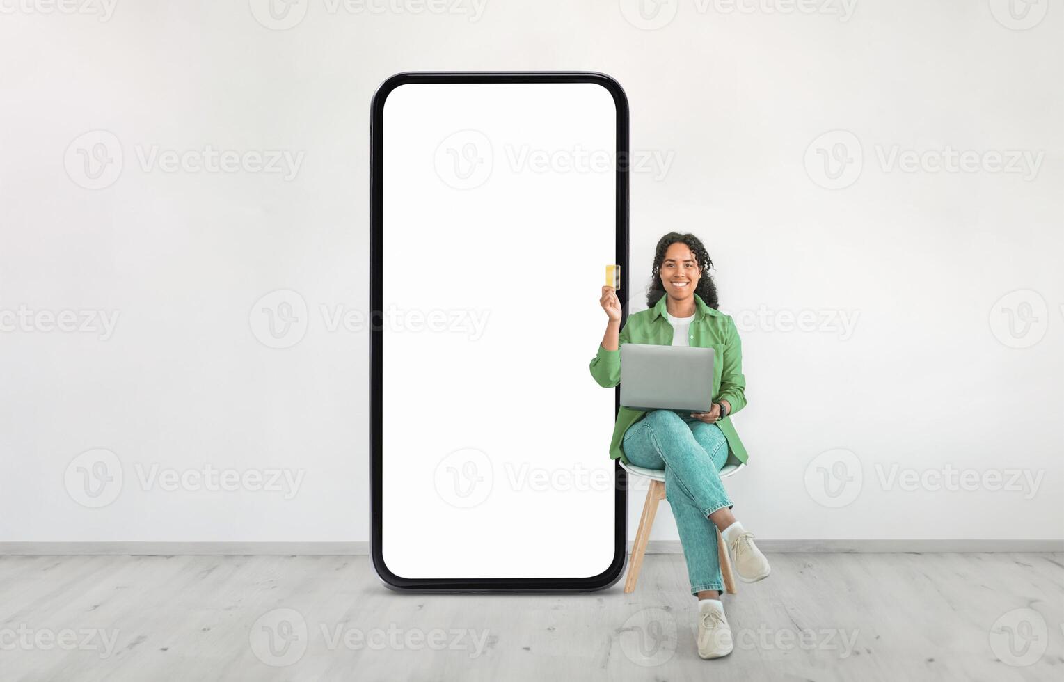 A woman sits on a chair, smiling as she works on a laptop. Beside her is an oversized blank screen, creating a modern and minimalist workspace environment. photo