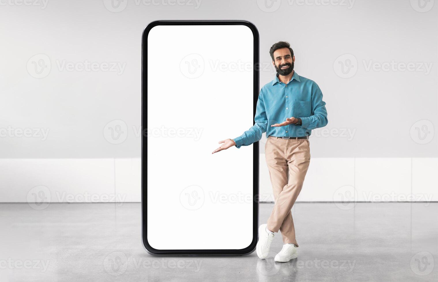 Middle eastern man in a blue shirt and beige pants stands next to a large blank smartphone screen, gesturing as if inviting viewers to engage with the display in a contemporary setting. photo