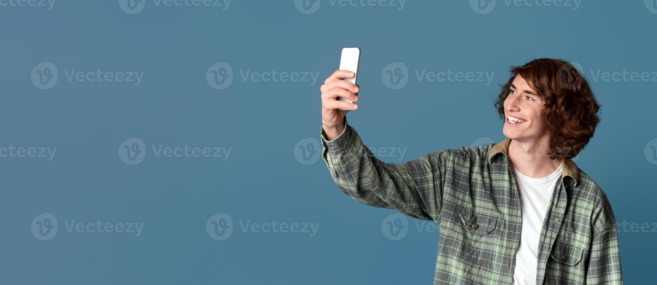 Curly teenage guy with smartphone taking selfie on turquoise background photo