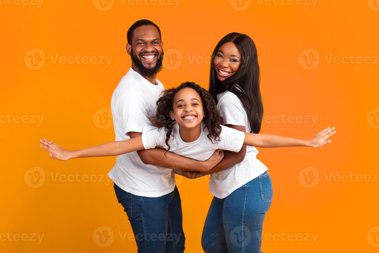 Bonding. Portrait of African American man and woman holding daughter on hands, standing isolated on yellow studio wall. Smiling girl pretending she is flying, spreading hands, imitating plane photo
