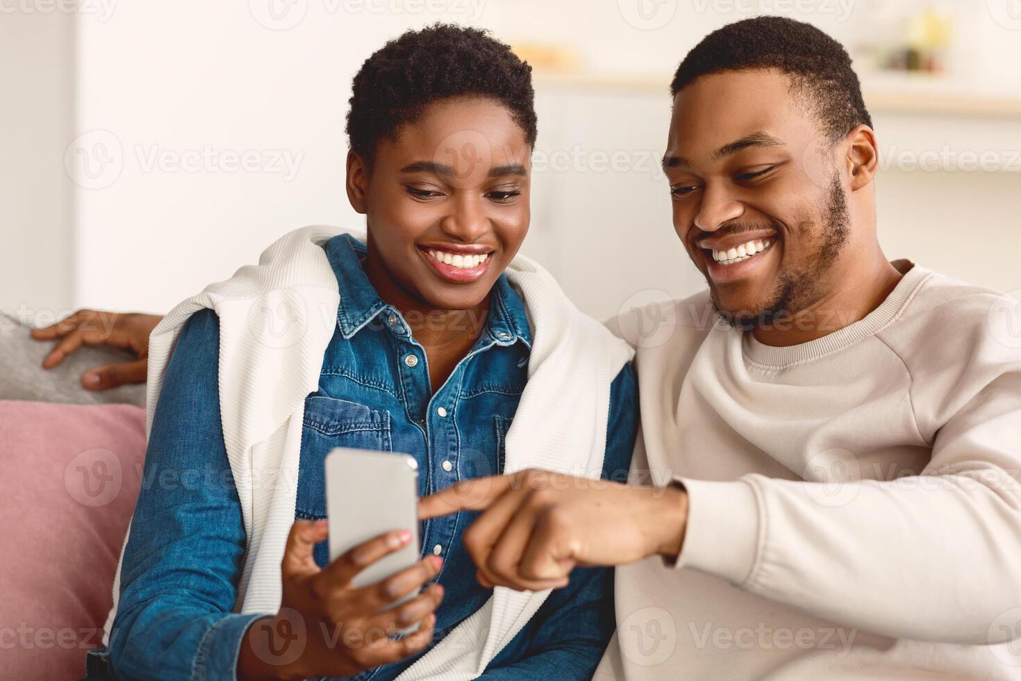 Portrait of positive young African American couple sitting on the sofa, using smartphone. Smiling woman holding her gadget, bearded man pointing at screen, showing photos, sharing social media content photo