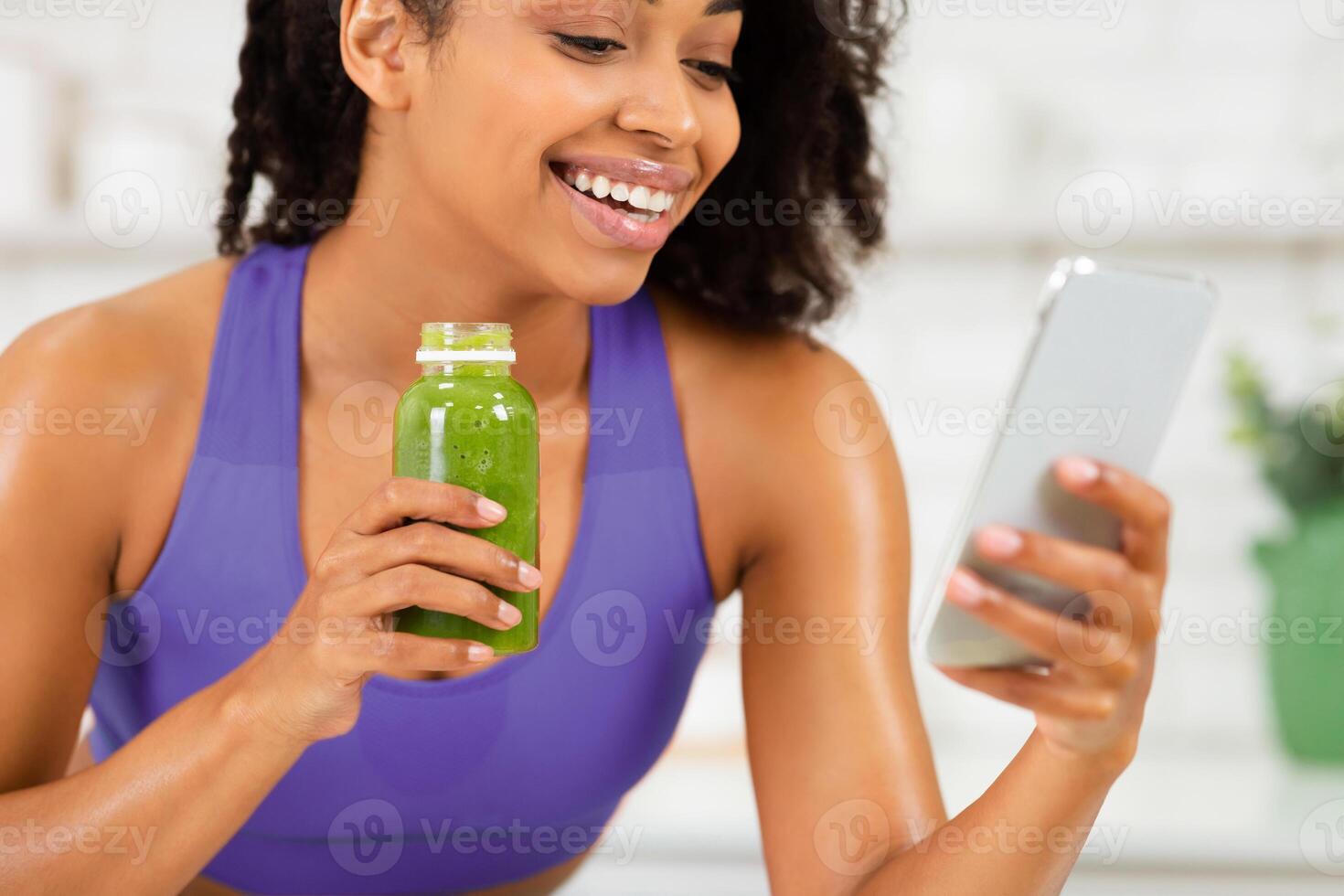 Surfing Internet. African girl in sportive clothing using smart phone and holding homemade smoothie in kitchen photo