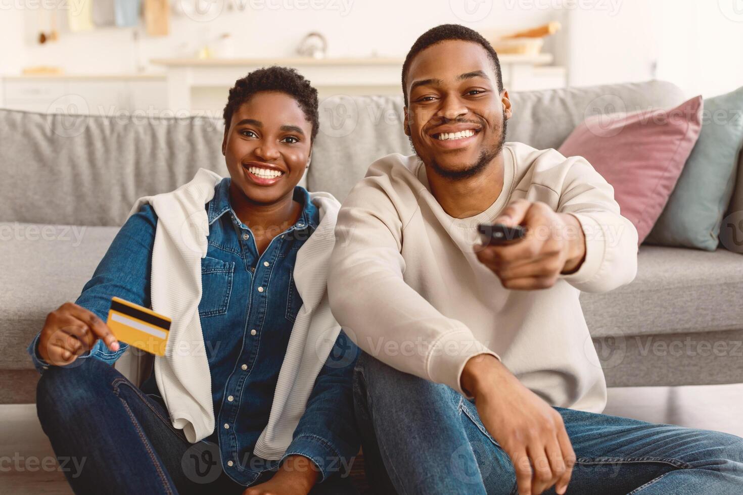 On Demand Concept. Smiling African American millennial couple sitting on floor, guy using tv remote controller, lady holding golden credit card, paying for vod service, multimedia streaming photo