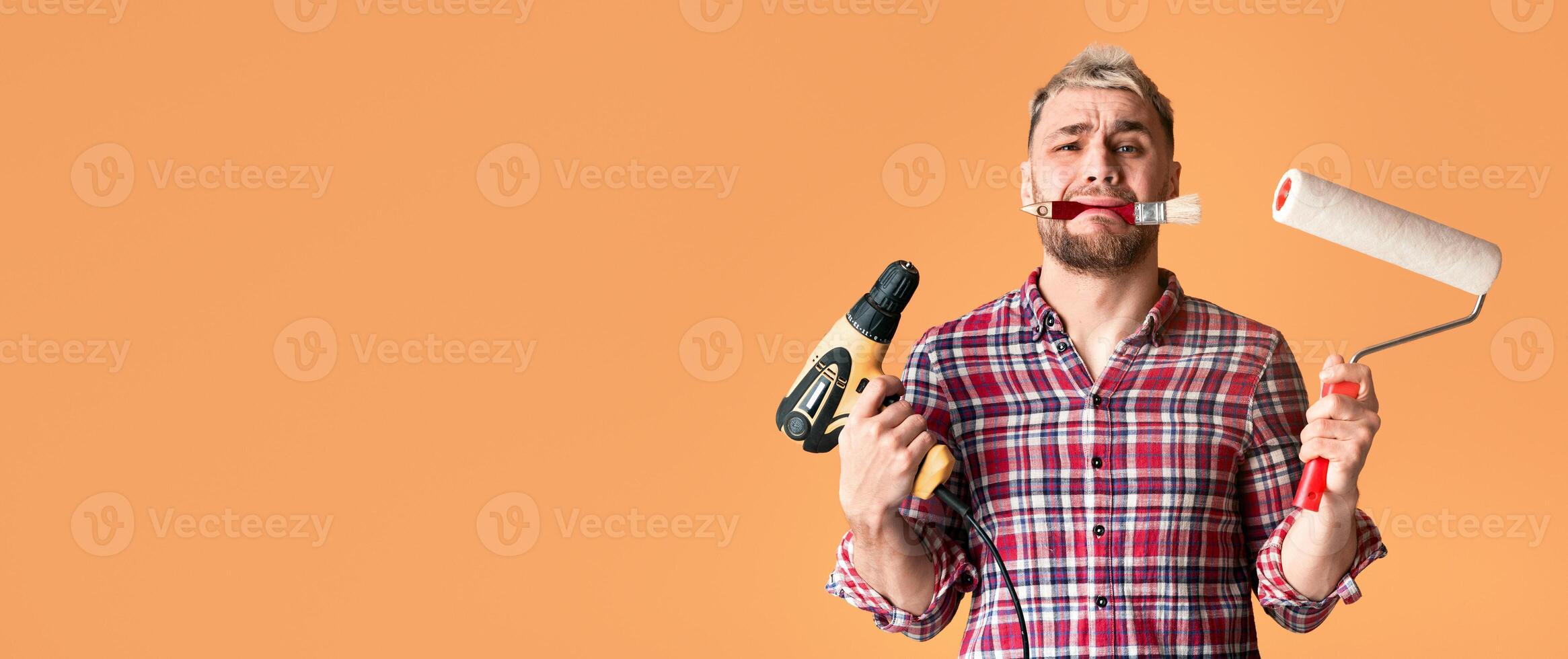 Multitasking problem. Desperate repairman holding perforator, paint roller and brush simultaneously, yellow studio background photo