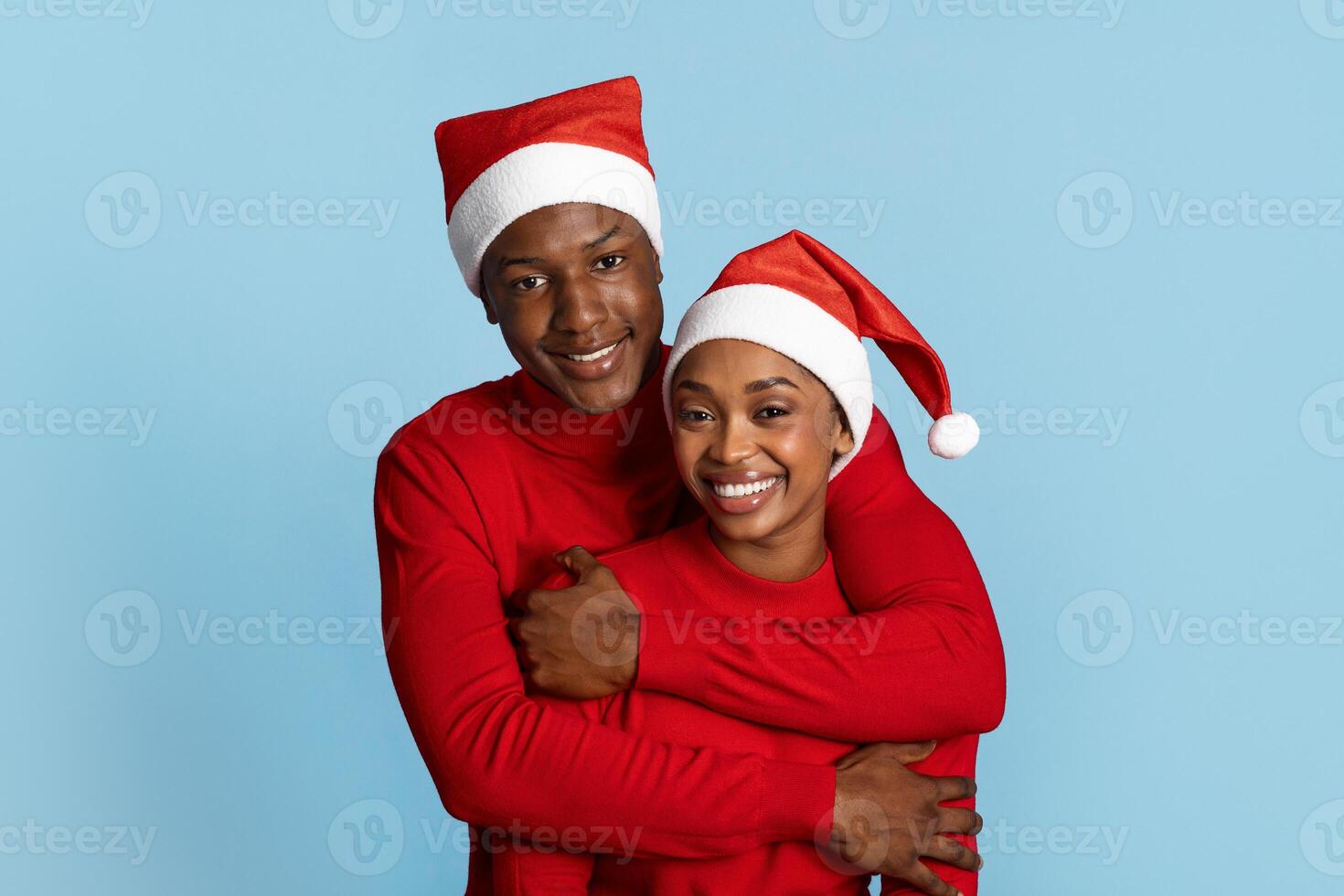 A joyful black couple embraces against a blue background, both wearing matching red sweaters and festive Santa hats. They radiate happiness and togetherness during the holiday season. photo