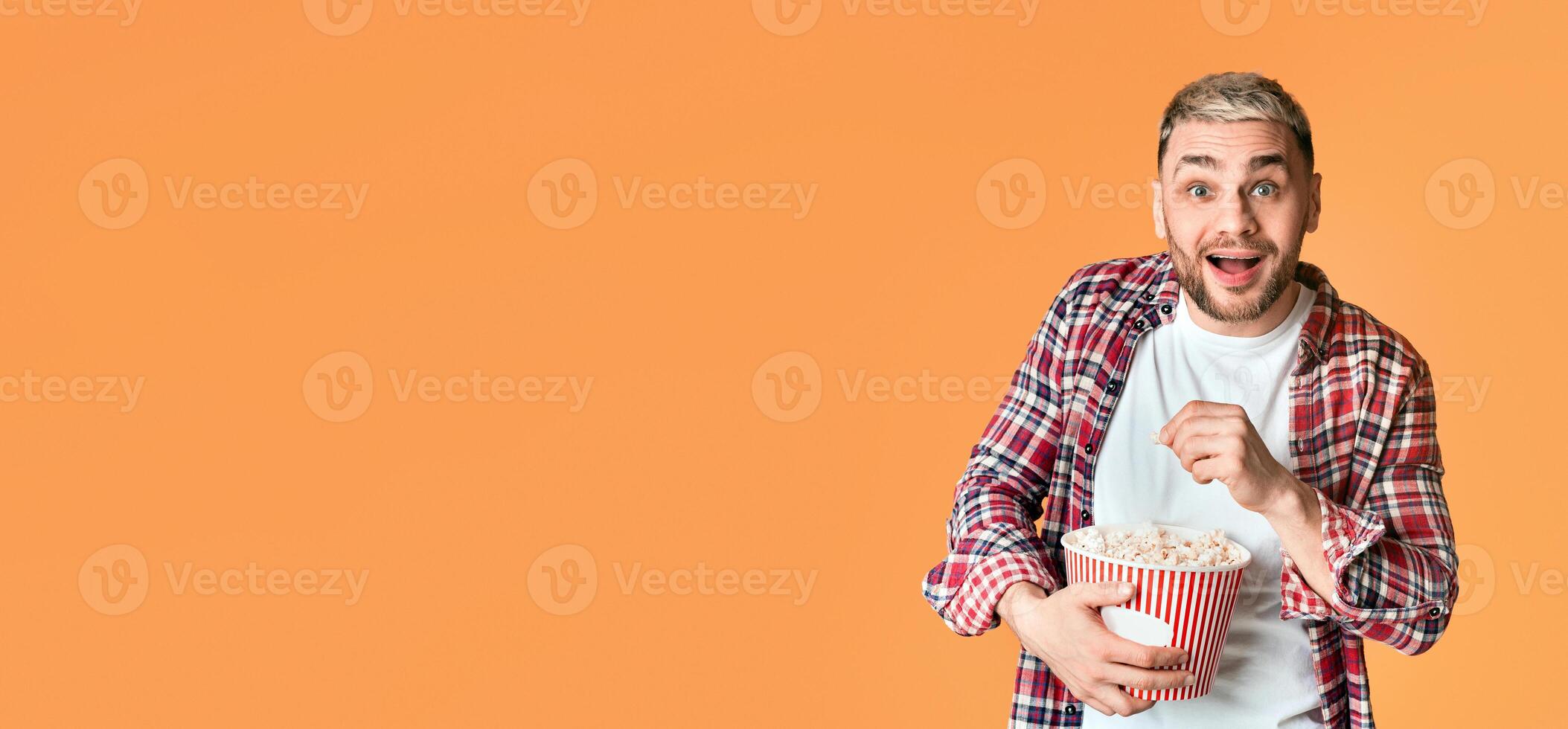 Impressed millennial guy watching movie emotionally and eating popcorn on yellow background, empty space photo