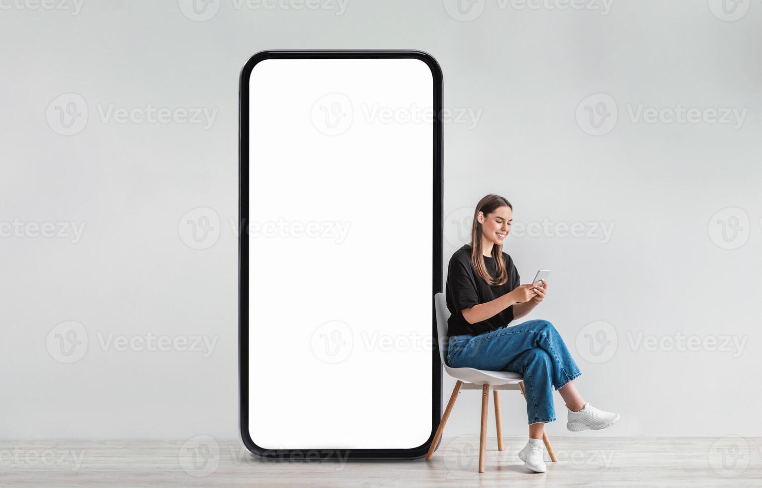 A young woman sits comfortably on a chair, engaging with her smartphone while a large blank screen stands next to her in a contemporary space, suggesting a digital connection. photo