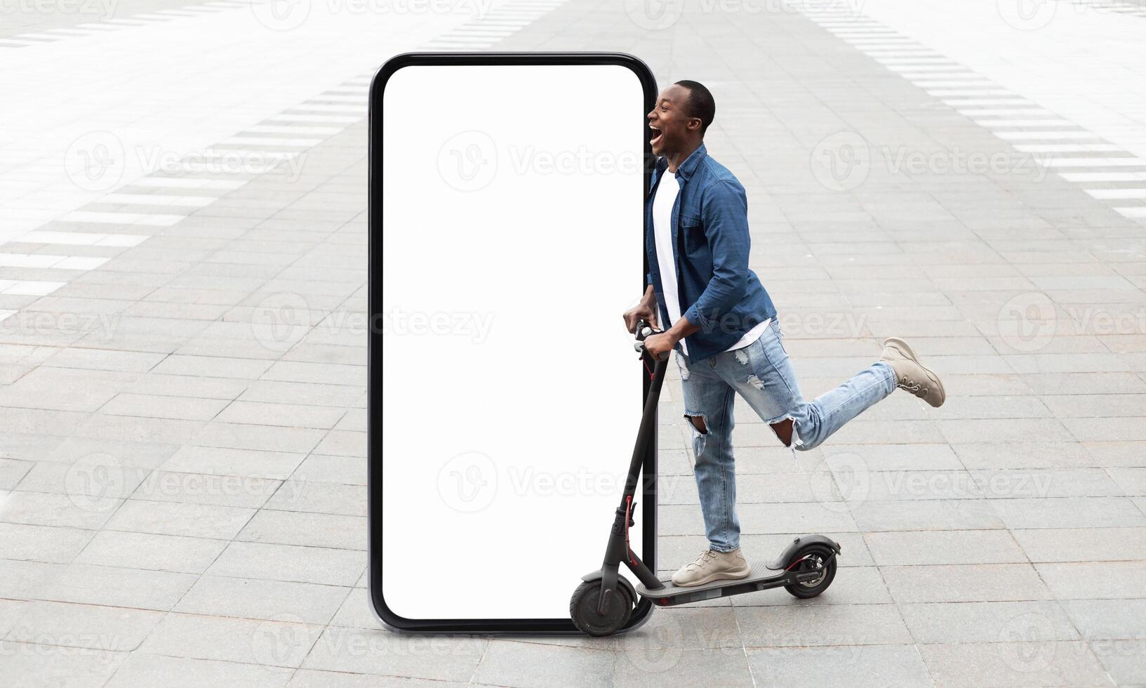 A young black man joyfully rides an electric scooter, playfully posing next to a large blank phone screen set in an urban area. The scene captures a moment of fun and creativity. photo