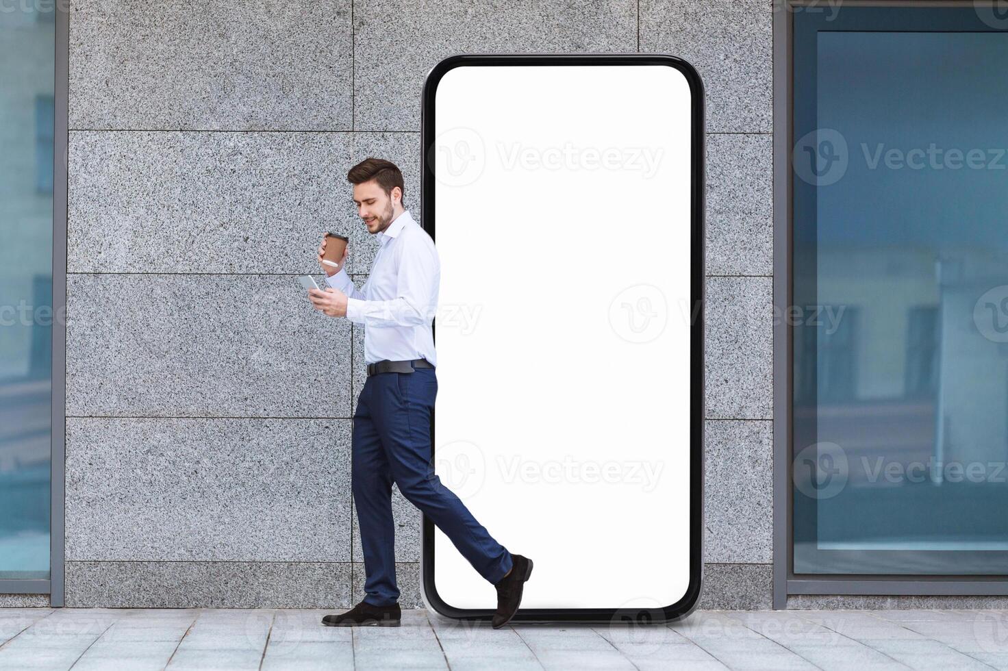 A man dressed in smart attire strides past an oversized blank digital screen on a city sidewalk, holding a coffee cup in one hand and checking his phone with the other. photo