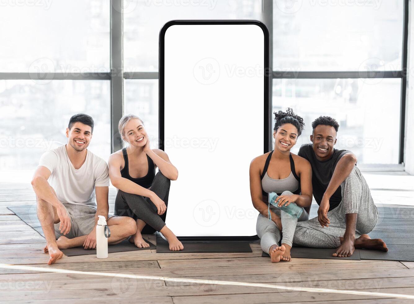 A group of four friends relaxes in a spacious, well-lit yoga studio, seated on the floor next to a blank backdrop. They share smiles and camaraderie after a session of physical activity. photo