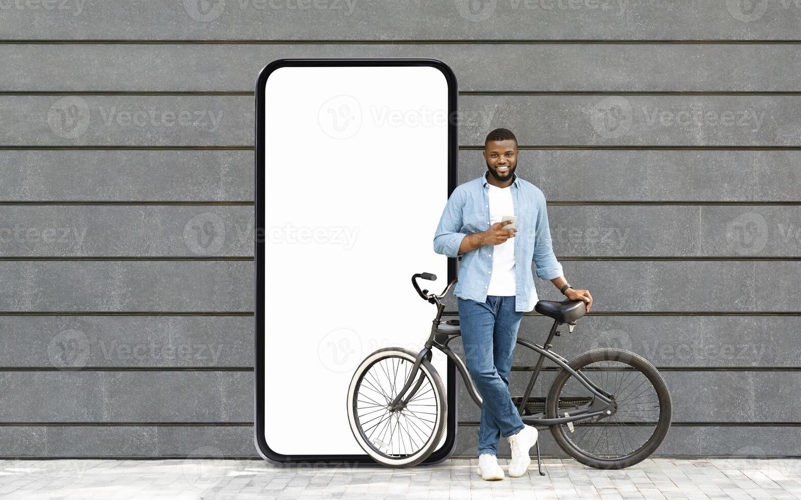 A young black man stands casually with a smartphone in hand, leaning against his bike, next to a large blank screen resembling a smartphone, set against a modern wall. photo