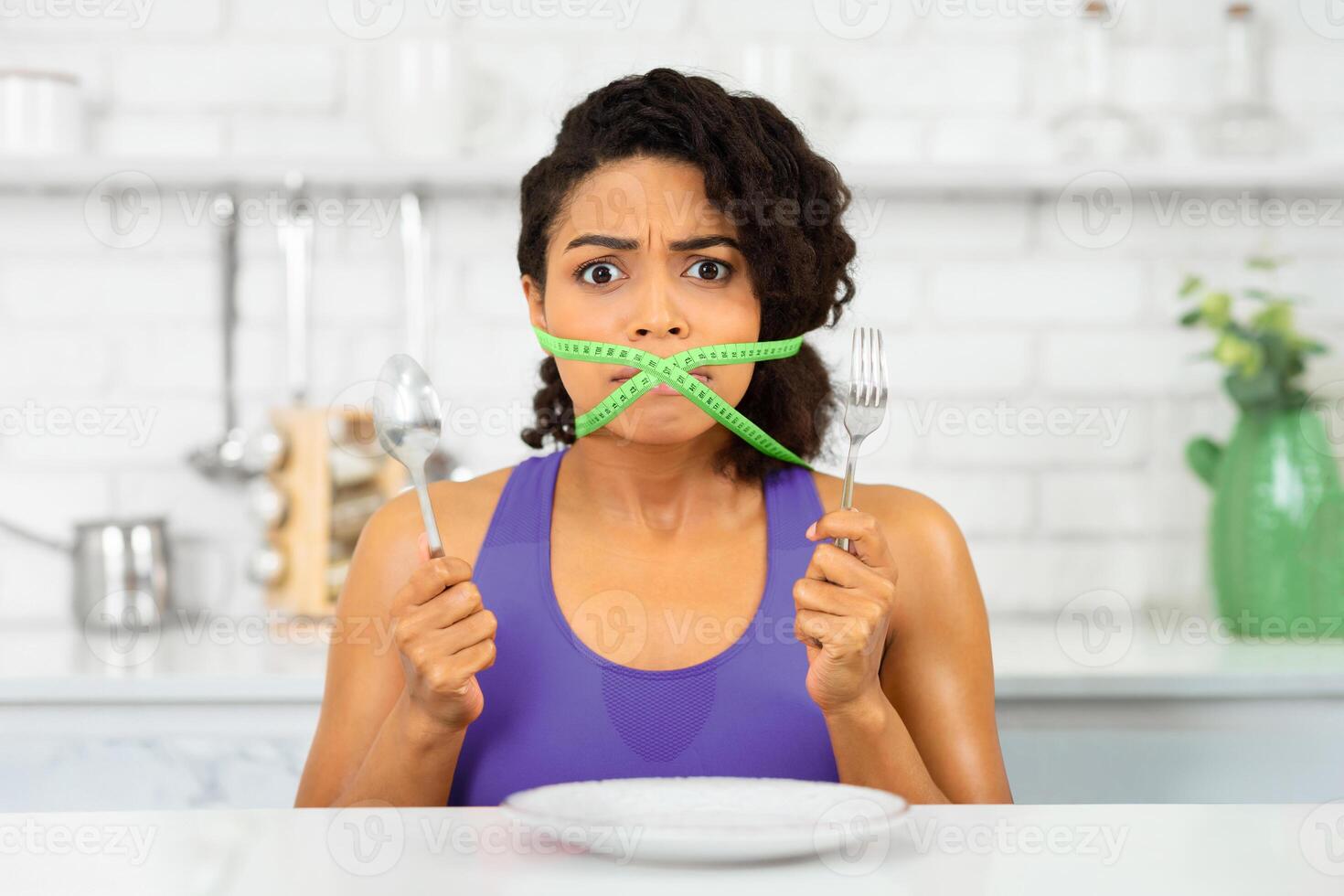 Diet Restriction Concept. Portrait of young frustrated black girl with a green measuring tape around her mouth photo