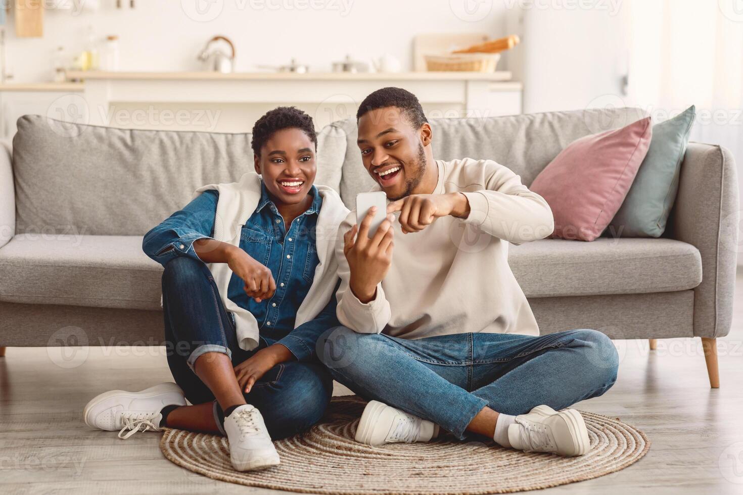 Portrait of positive young African American couple sitting on the floor carpet, using smartphone. Smiling man holding his gadget and pointing at screen, showing photos, sharing social media content photo