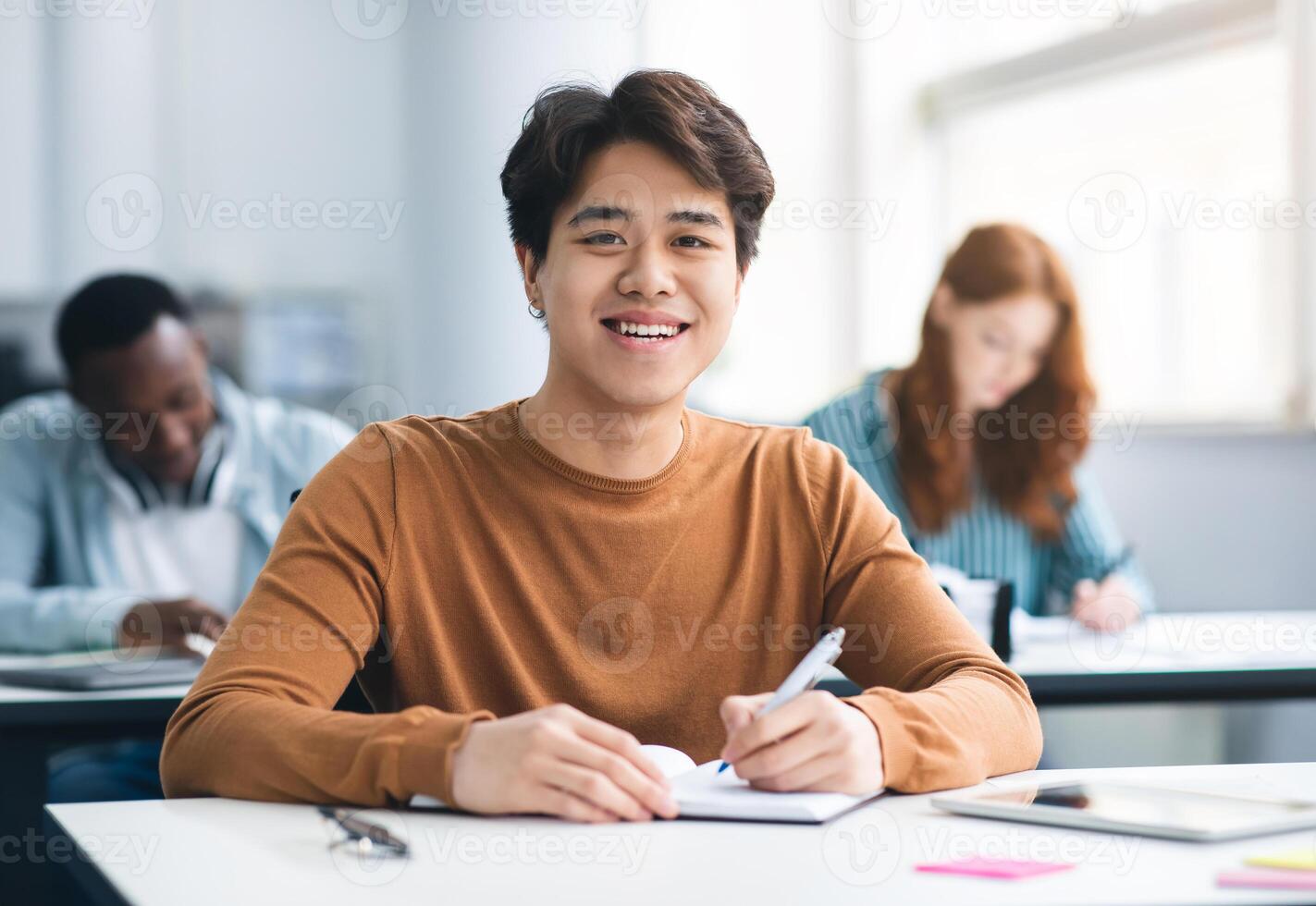 Modern Education Concept. Portrait of smiling asian male student sitting at desk in classroom at university, taking test or writing notes in his notebook, looking posing at camera, selective focus photo