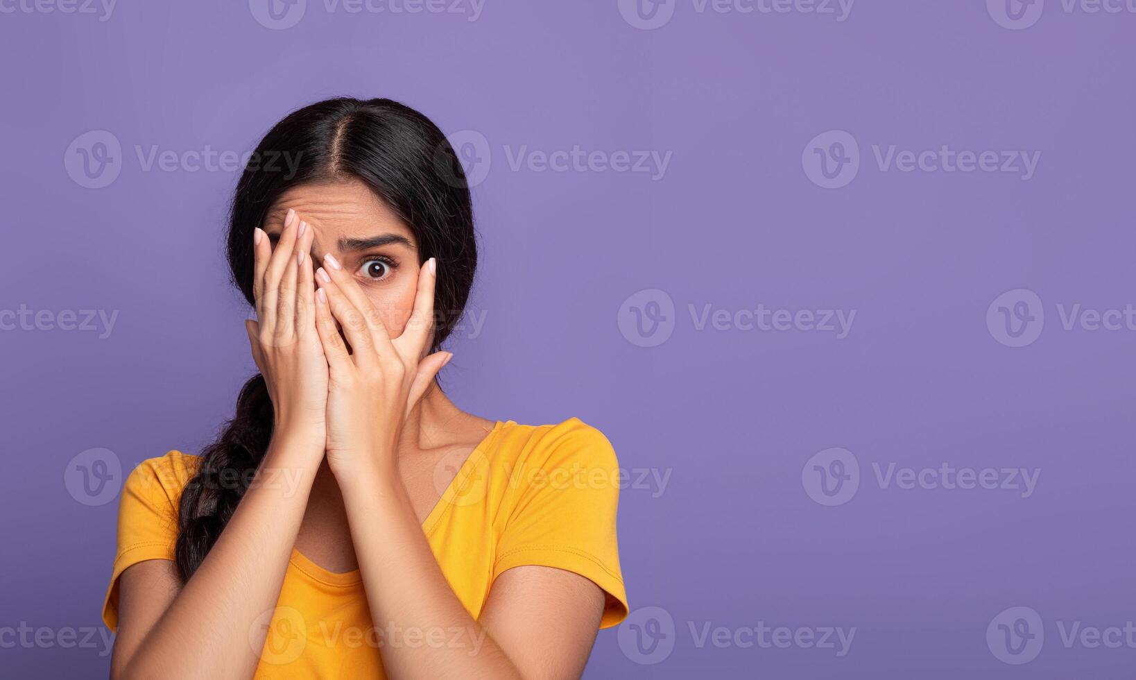 Portrait of shocked and scared young indian lady covering her eyes with palms and peeking through fingers, standing isolated over purple studio background, banner, free copy space. Phobia Concept photo