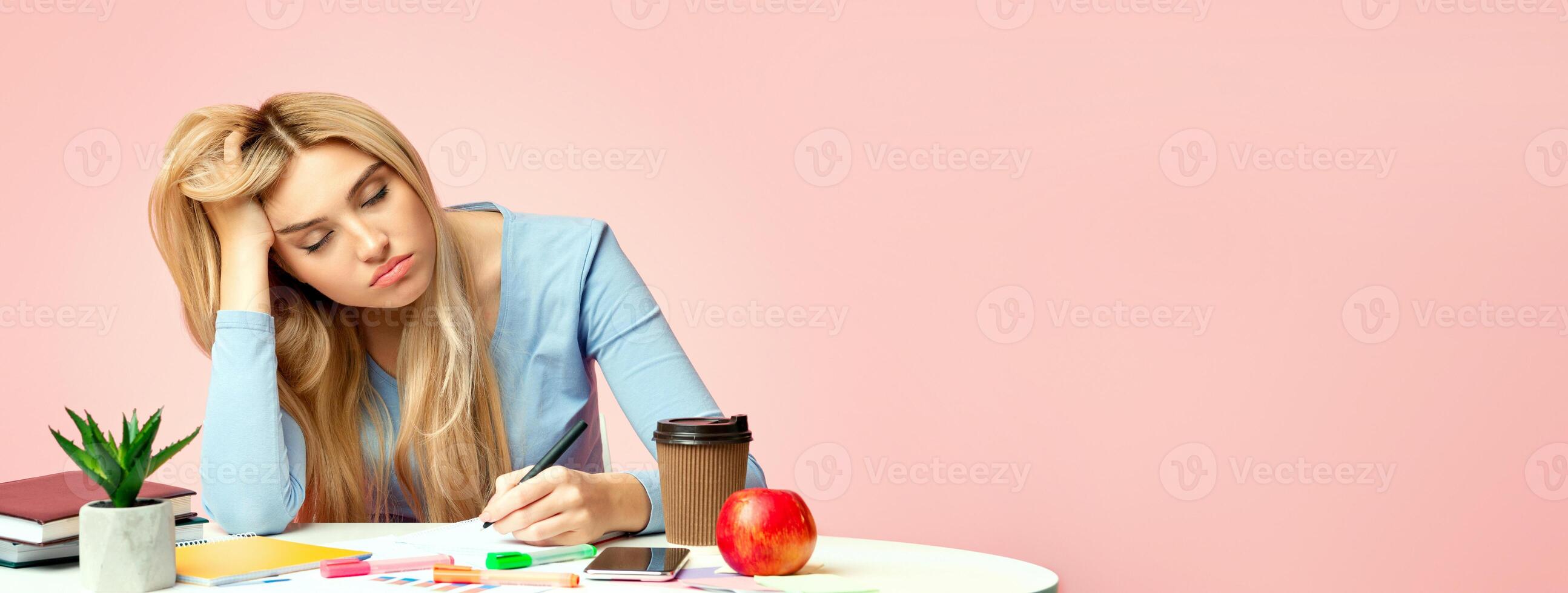 Sleep At Work. Portrait of sad blonde student sitting at table resting head on hand and writing, copyspace photo